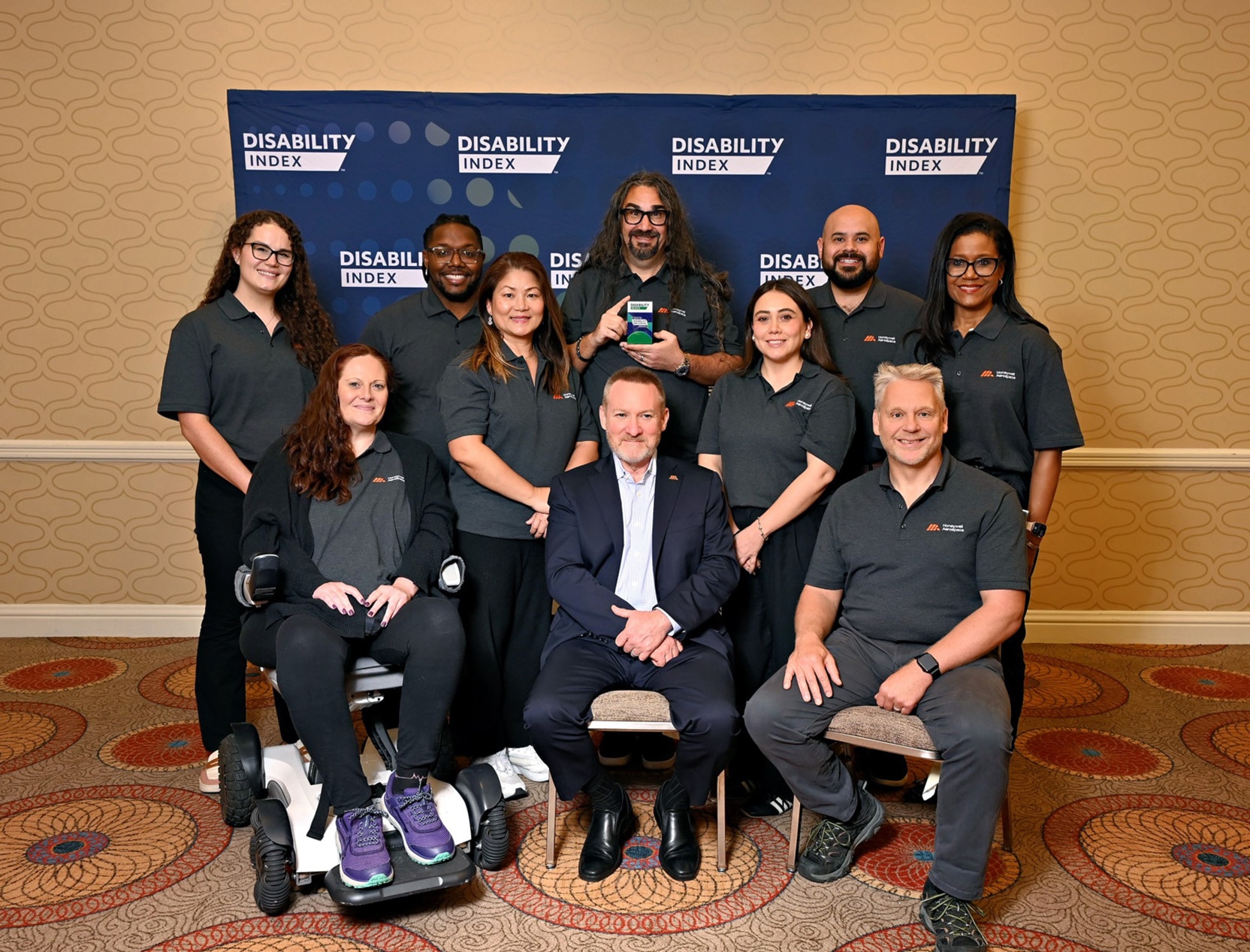 Honeywell Aerospace team members gather in front of a Disability Index backdrop, standing and sitting together, including one person using a mobility device. Wearing matching gray shirts, the group smiles in a conference room with patterned carpet and neutral walls. The photo reflects Honeywell Aerospace’s commitment to inclusion, accessibility, and a supportive workplace culture.
