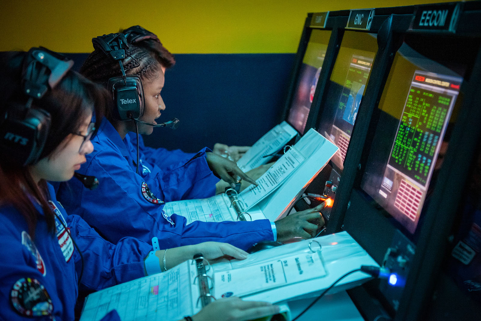 Students participate in a mission control simulation, wearing headsets and reviewing binders while monitoring data on multiple screens.