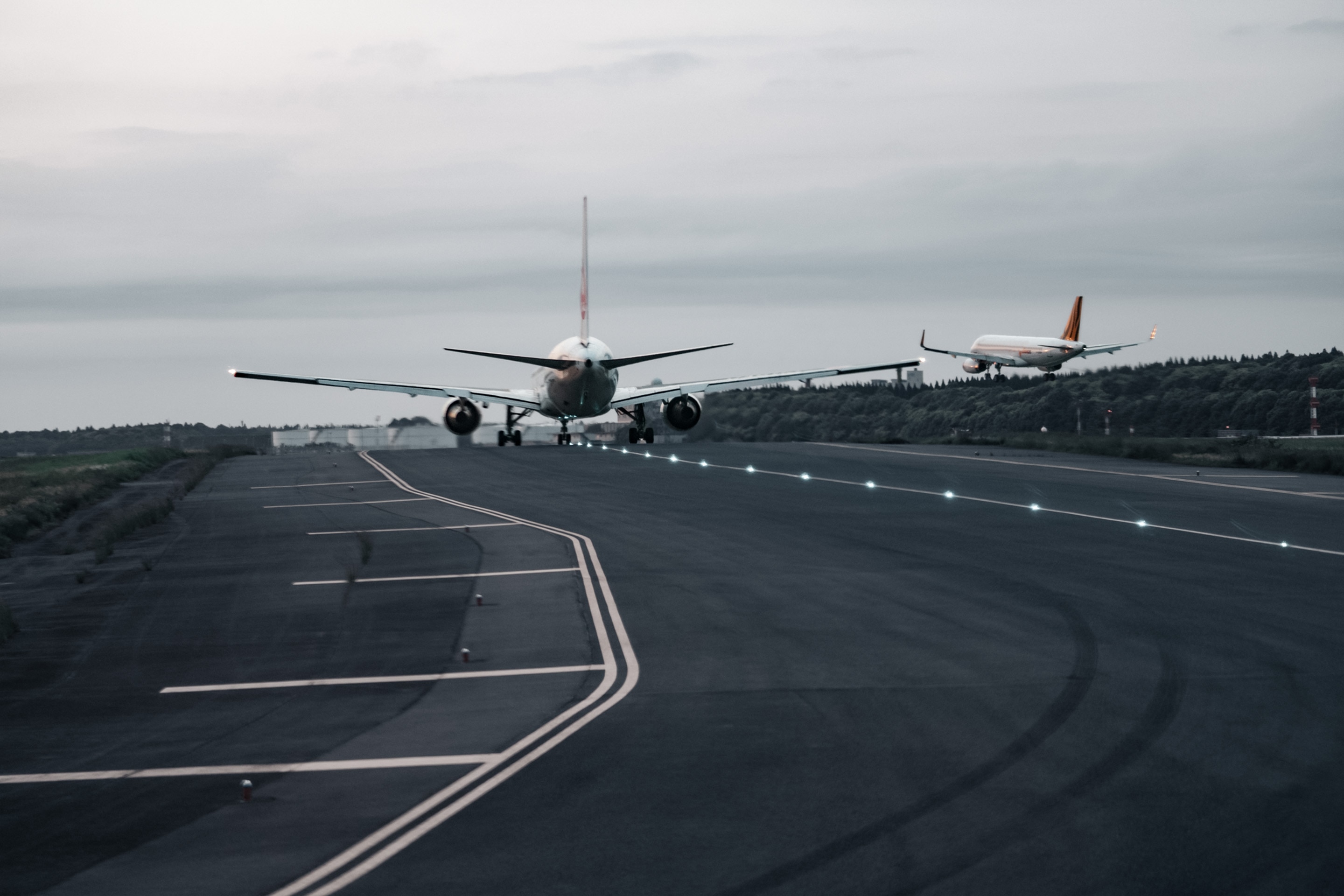 Rear view of a jet taxiing on a runway with another aircraft approaching, runway lights and markings leading into a cloudy horizon