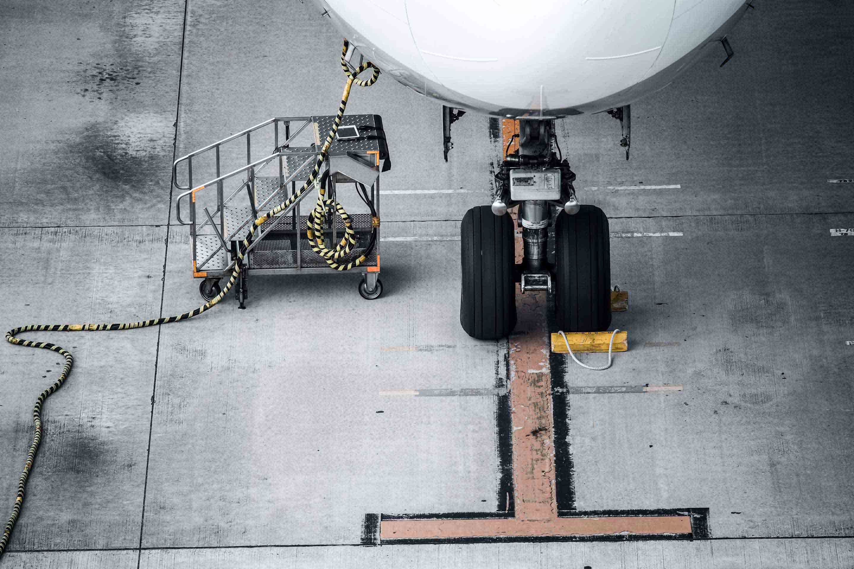 Overhead aerial view of a commercial aircraft nose landing gear parked on a hangar floor with yellow wheel chocks and a hose coiled nearby, showing the nose gear strut, torque links, and twin wheels from directly above. This image highlights Honeywell's landing gear systems.