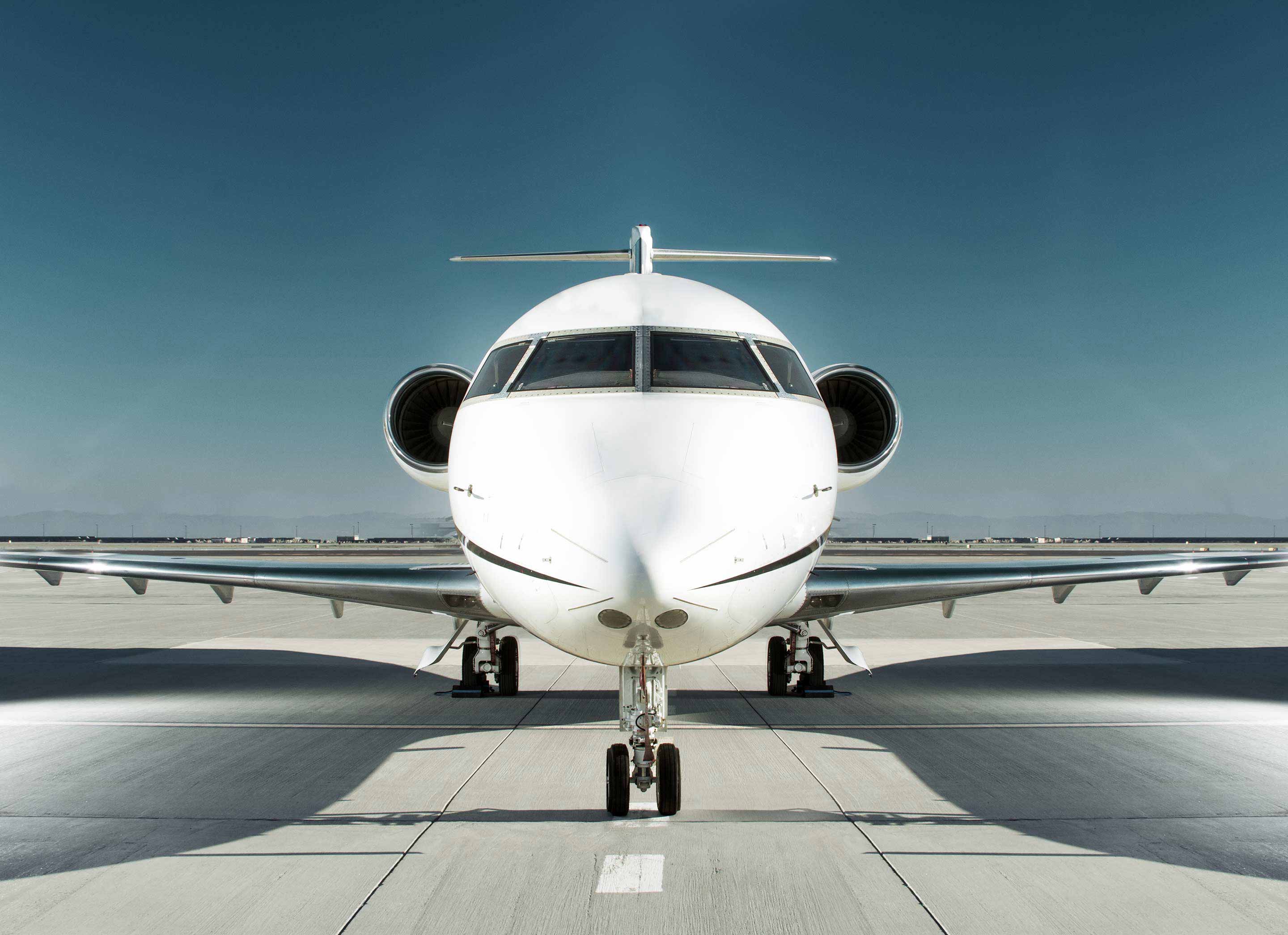 Perfectly centered head-on photograph of a sleek white business jet parked on a tarmac with twin wing-mounted engines clearly visible and a deep blue sky above. This clean marketing-style image represents Honeywell's business jet avionics, APU, and propulsion solutions.