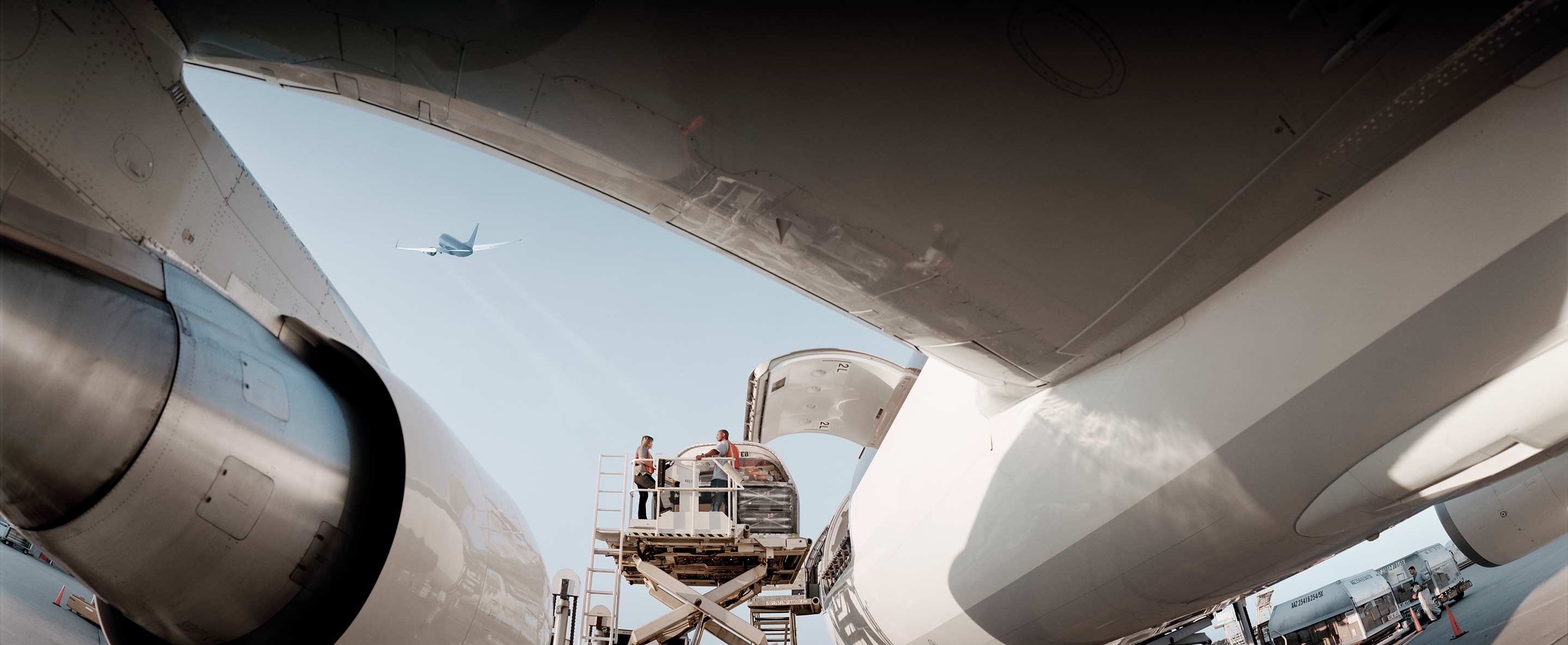 Wide-angle perspective shot from between two wide-body aircraft fuselages looking up at maintenance workers on a platform, with a departing aircraft visible between the aircraft noses. This image represents Honeywell's aircraft MRO, engine servicing, and APU solutions.