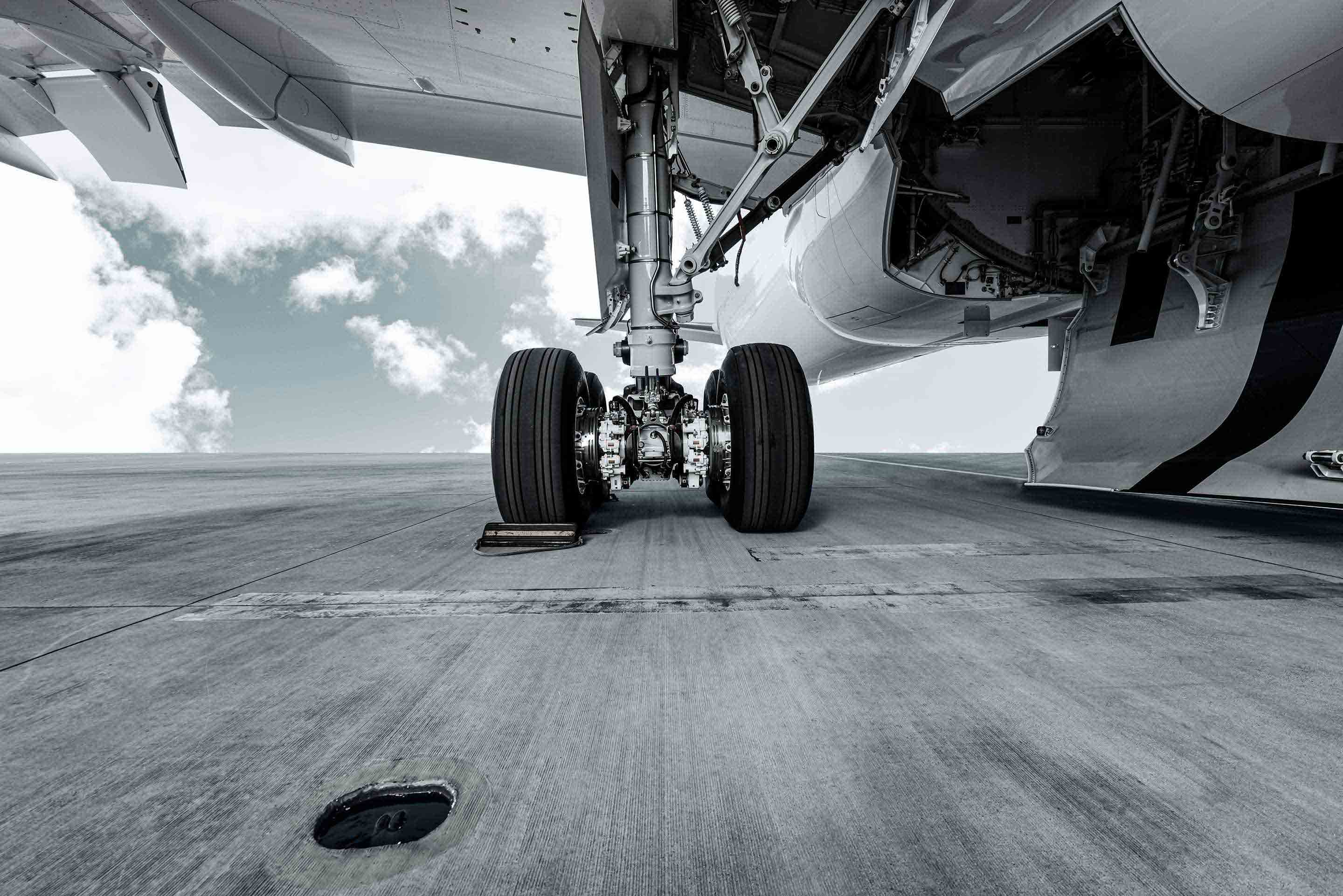 Ground-level worm's-eye view of a commercial aircraft's nose landing gear assembly parked on a tarmac, with the fuselage belly and clouds above dramatically framed against a blue sky. This image highlights Honeywell's landing systems and aircraft wheel and brake technology.