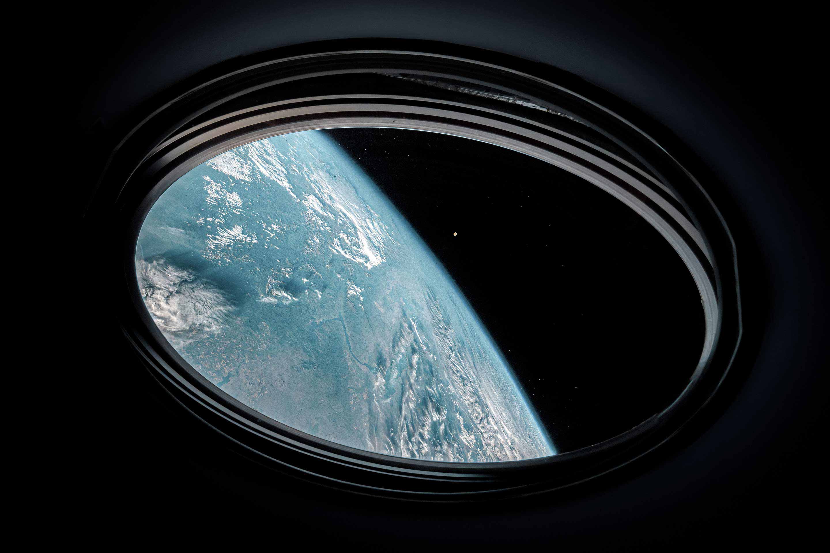 Stunning view of Earth's curvature and cloud patterns seen through a large circular porthole or viewing window of a space station in low Earth orbit, with space visible at the edges. This space technology image reflects Honeywell's space systems and satellite navigation capabilities.