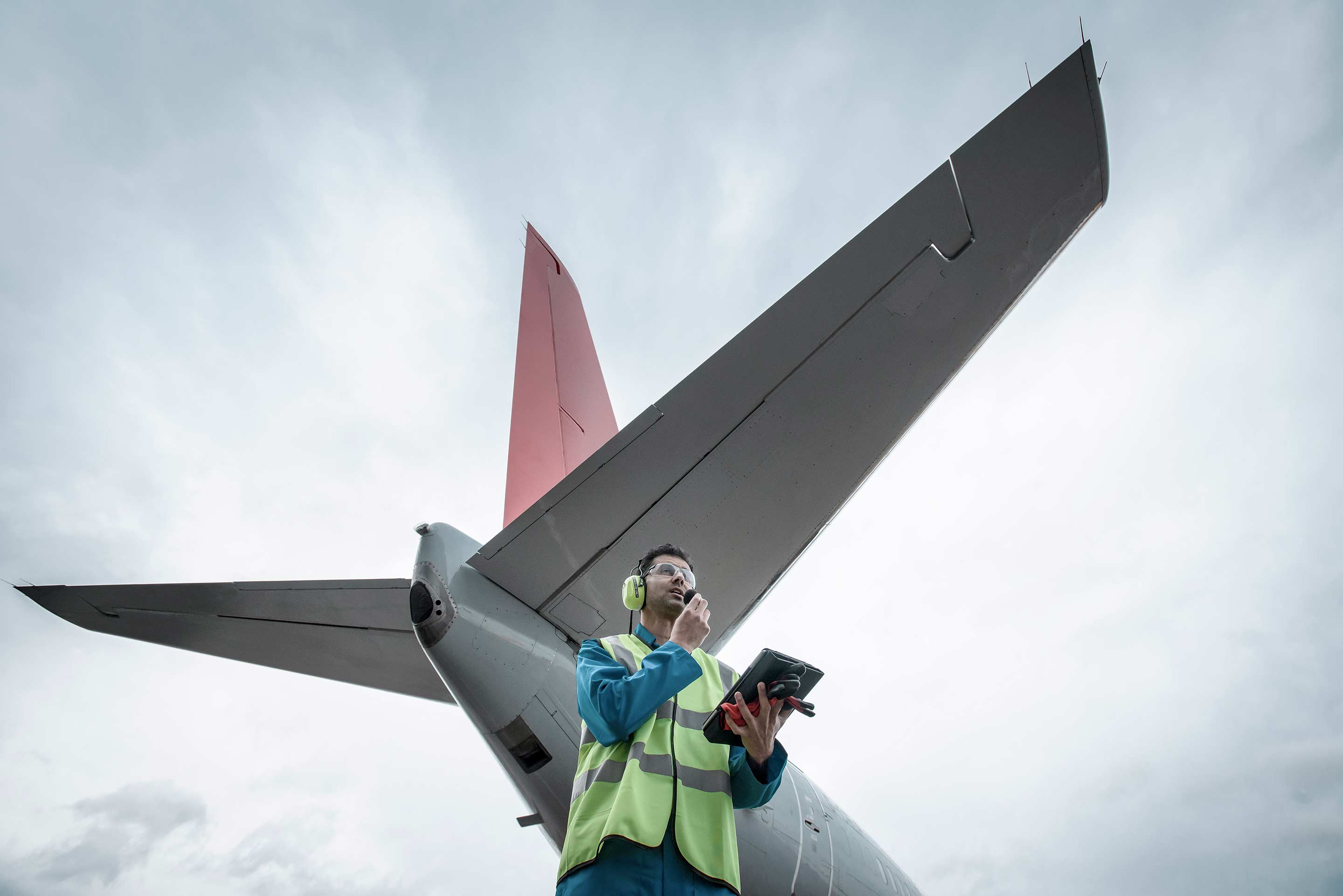 An aviation maintenance worker in a safety vest stands beneath the tail section of a red-tailed commercial aircraft, speaking on a radio while holding a tablet. This image represents Honeywell's digital MRO, Forge analytics, and connected aircraft maintenance platform.