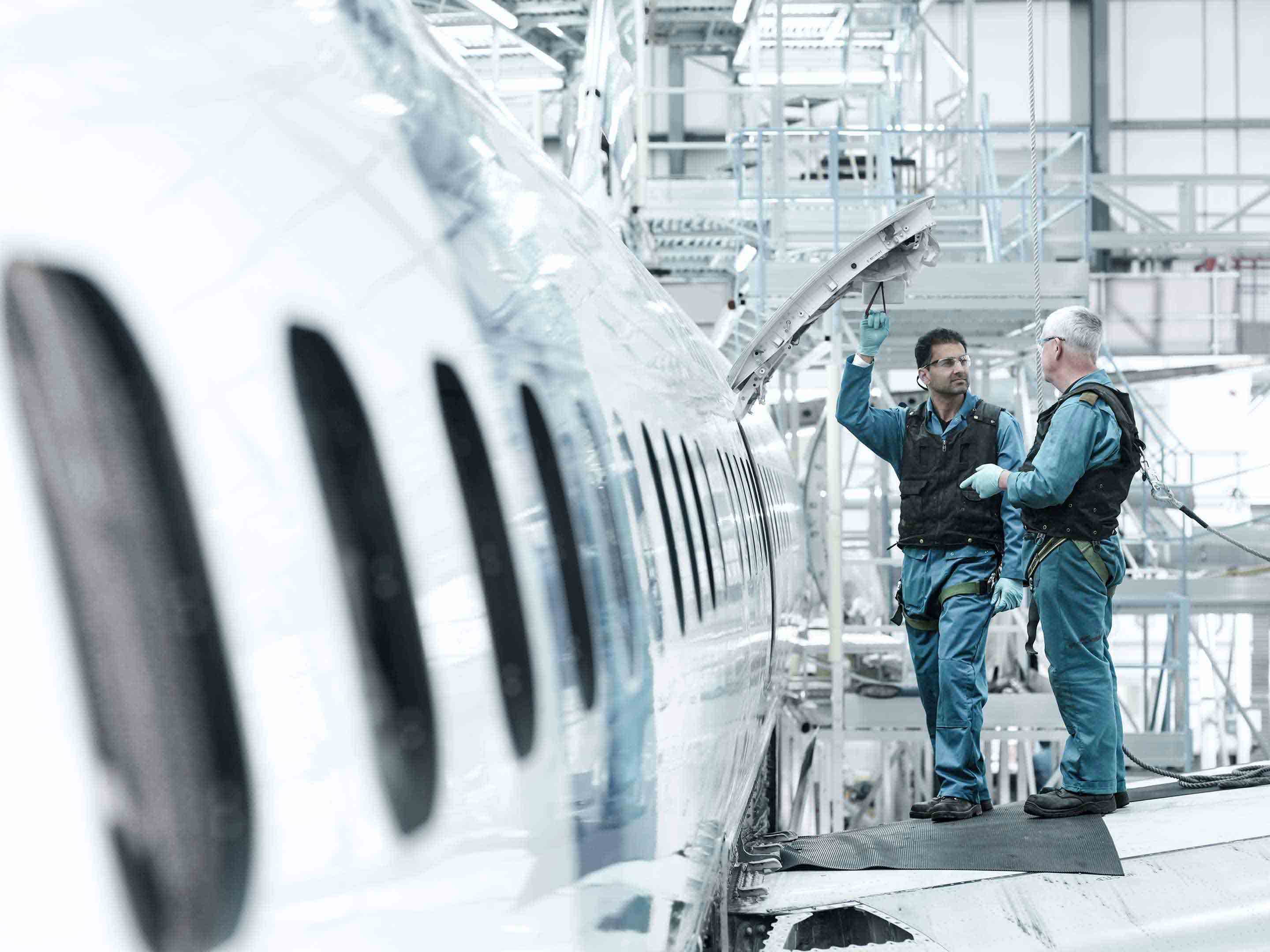 Two aircraft maintenance technicians in dark overalls open an access panel on a business jet fuselage in a bright MRO hangar, with one pointing to an item inside. This hands-on MRO image reflects Honeywell's business jet maintenance and aftermarket support services.