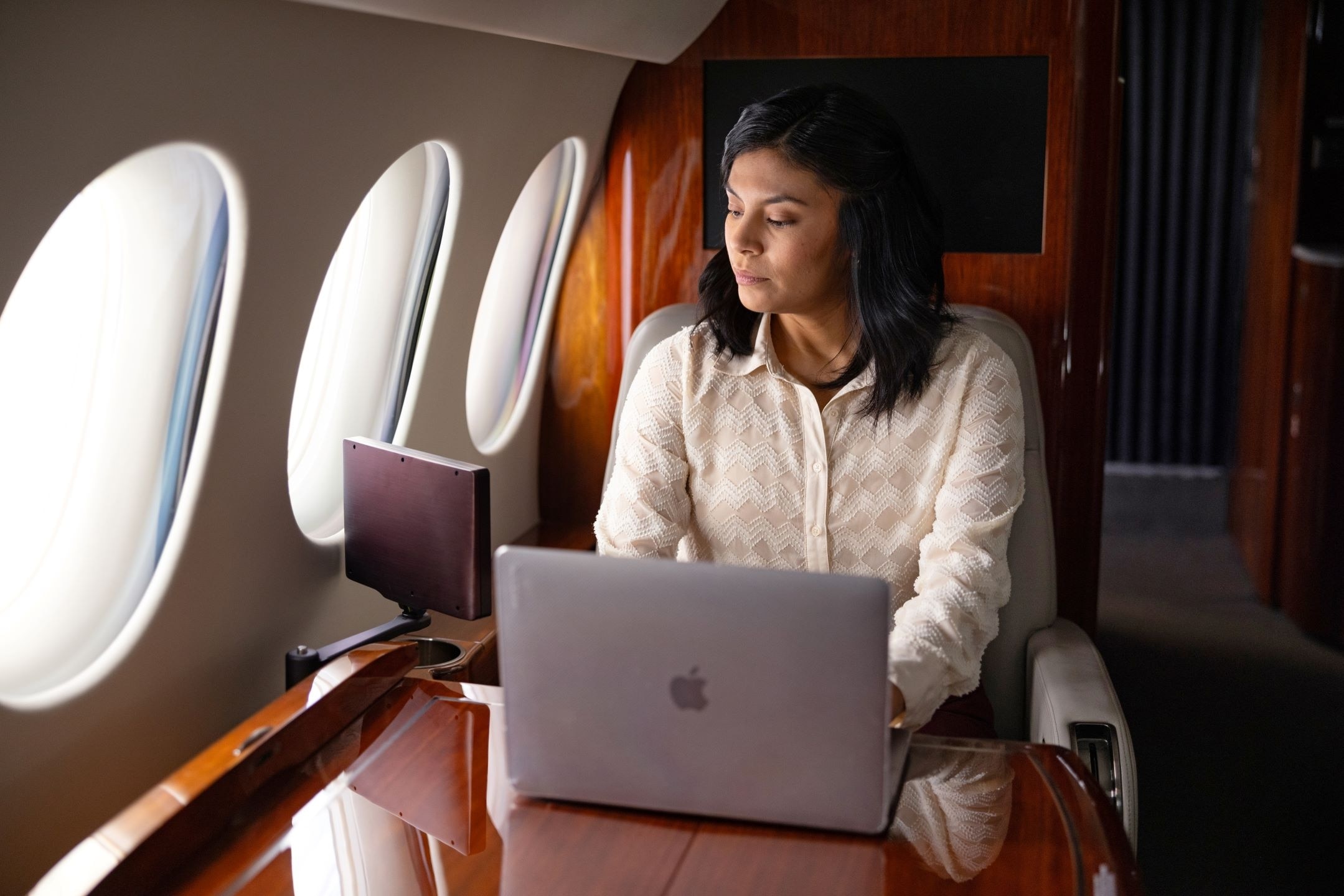 A person working on a laptop in a luxurious jet cabin with wood paneling, oval windows, and a polished wooden table.